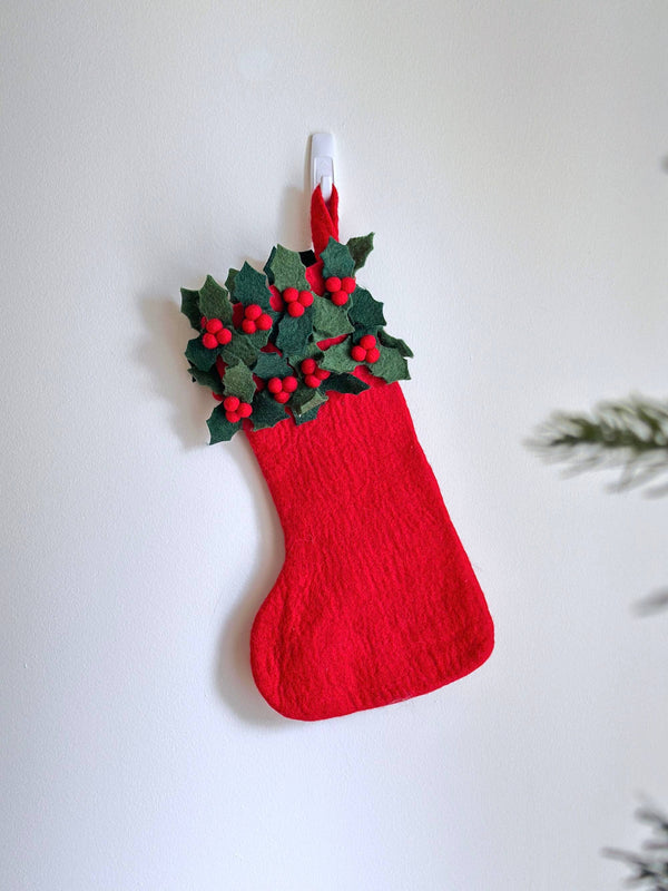 A vibrant Felt Holly Christmas Stocking hangs from a clear hook against a bright white wall, evoking a joyful and festive mood. The textured red stocking is adorned with clusters of green holly leaves and small red berries at the top. A Christmas tree branch is partially visible in the lower right.