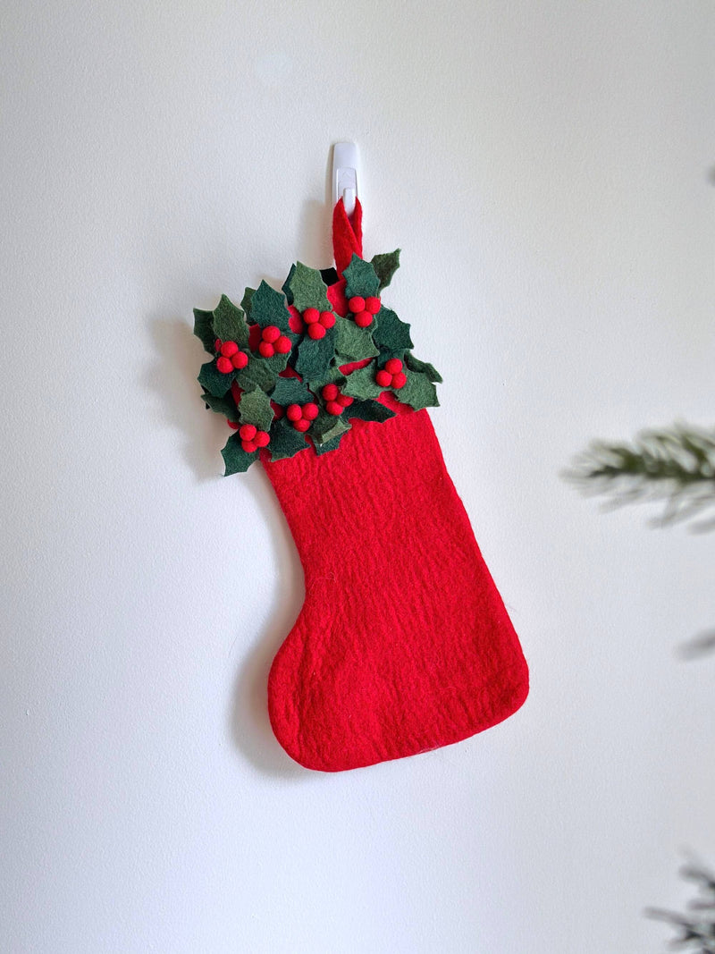 A vibrant Felt Holly Christmas Stocking hangs from a clear hook against a bright white wall, evoking a joyful and festive mood. The textured red stocking is adorned with clusters of green holly leaves and small red berries at the top. A Christmas tree branch is partially visible in the lower right.