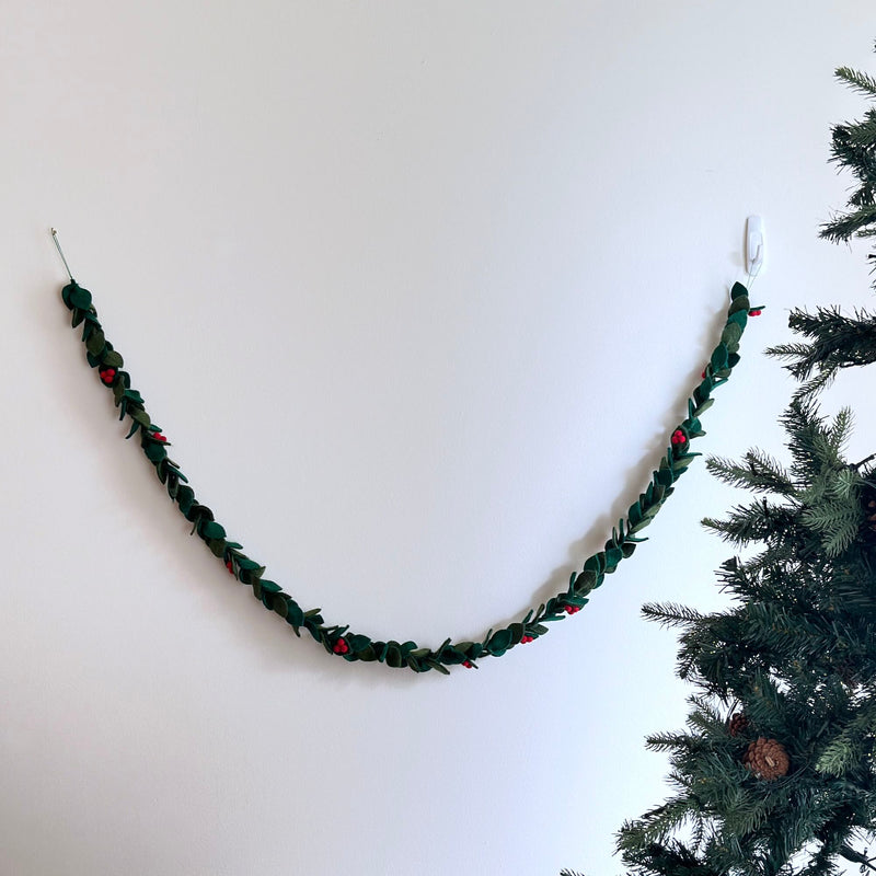 A wool felt holly garland hangs on a white wall next to a Christmas tree, creating a festive atmosphere. The garland features alternating dark green whimsical holly leaves and small red berries strung along a jute thread, forming a cheerful pattern.