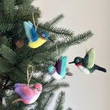 Four needle felted hummingbird Christmas ornaments hang from the branches of a lush green Christmas tree. Each wool felt hummingbird features a slender black beak, small wings, and a jute string for hanging, adding a charming touch to the festive decor.