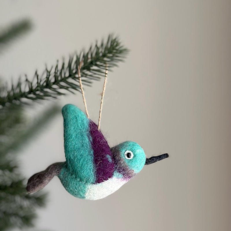 A needle felted Felt Hummingbird Christmas ornament is displayed on a Christmas tree branch, evoking natural beauty. This 100% wool felt hummingbird features a harmonious blend of colors on its wings, body, and head. A thin, black beak and a jute string for hanging complete its design.