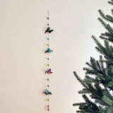 A colorful felt hummingbird garland is displayed against a plain wall, next to a Christmas tree. This handmade garland features four felt hummingbirds in vibrant combinations: green and red, light blue and pink, purple and pink, and blue and yellow. Each hummingbird is separated by felt balls in shades of gray, light blue, green, and purple, creating a cheerful decorative accent.