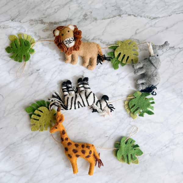 A handcrafted felted wool jungle animal garland is displayed on a white marble surface, presenting a playful and natural aesthetic. It features a tan lion with a dark brown mane, a gray elephant, a black and white striped zebra, and an orange giraffe with brown spots. Green monstera leaves are interspersed among the animals, all connected by a rustic jute thread.