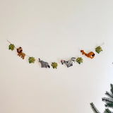 A wool felt jungle animal garland, featuring natural felted wool figures, is displayed against a plain white wall. The playful garland showcases a brown lion, a gray elephant, a black and white striped zebra with pink ears, and a spotted giraffe, alternating with green monstera leaves along a rustic jute thread. A hint of a coniferous tree is visible in the lower right corner.