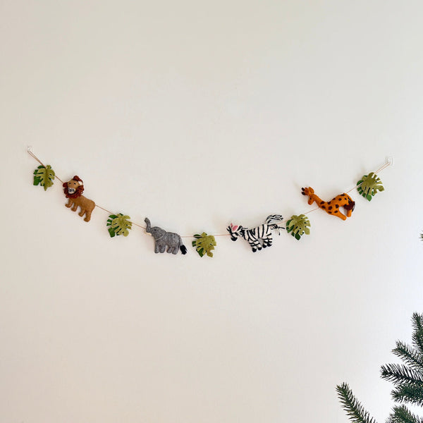 A wool felt jungle animal garland, featuring natural felted wool figures, is displayed against a plain white wall. The playful garland showcases a brown lion, a gray elephant, a black and white striped zebra with pink ears, and a spotted giraffe, alternating with green monstera leaves along a rustic jute thread. A hint of a coniferous tree is visible in the lower right corner.