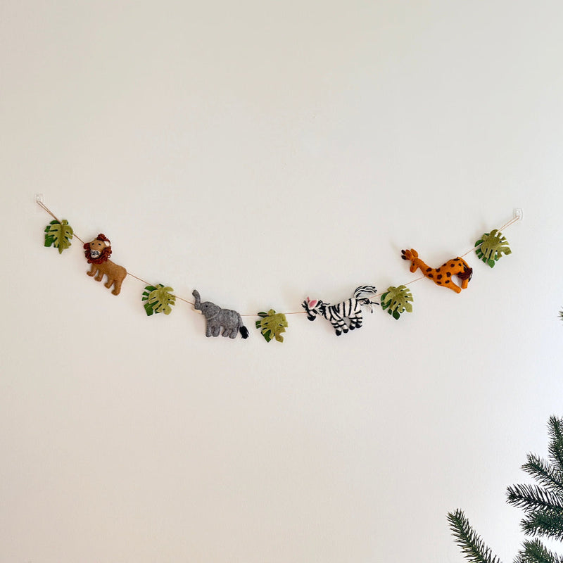 A wool felt jungle animal garland, featuring natural felted wool figures, is displayed against a plain white wall. The playful garland showcases a brown lion, a gray elephant, a black and white striped zebra with pink ears, and a spotted giraffe, alternating with green monstera leaves along a rustic jute thread. A hint of a coniferous tree is visible in the lower right corner.