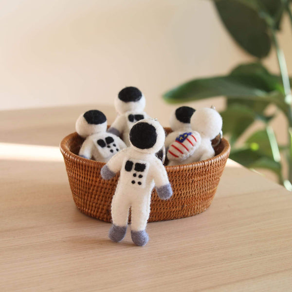 Wool felt astronaut keychains are artistically arranged inside and beside a woven brown basket, resting on a light wooden surface. These handcrafted keychains are primarily white, featuring black helmets, gray gloves and boots, and small dark square accents. One unique keychain also displays a tiny American flag, with each having a metal key ring attached.