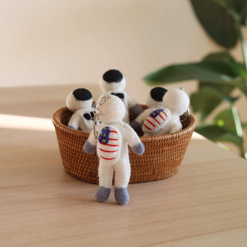 Several astronaut felt keychains are displayed on a light wood surface in a naturally lit setting, with one standing prominently in front of a woven basket holding more. Each felt astronaut keychain features a white body, gray hands and feet, and a black helmet. A metal keychain with key ring is attached to the front astronaut.