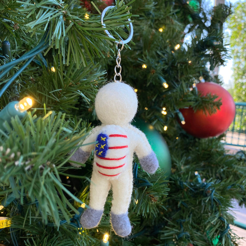 A wool felt astronaut keychain hangs on a festive Christmas tree, surrounded by soft twinkling lights and ornaments. This unique handmade felt keychain is primarily white with gray hands and feet. Its body displays a design resembling the American flag, featuring red stripes and a blue square with yellow stars, attached to a meta keychain.