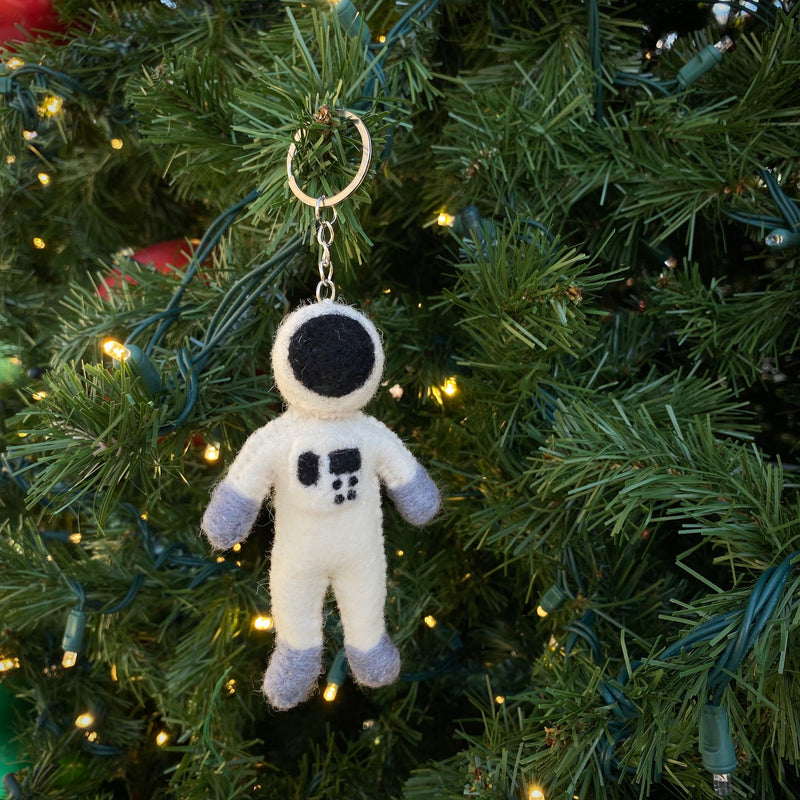 A handcrafted wool felt astronaut keychain hangs playfully on a lush, green Christmas tree, which is adorned with warm white lights. The stylized white astronaut features a black visor, gray hands and feet, and small black squares on its body. It is attached to a metal keychain with key ring.