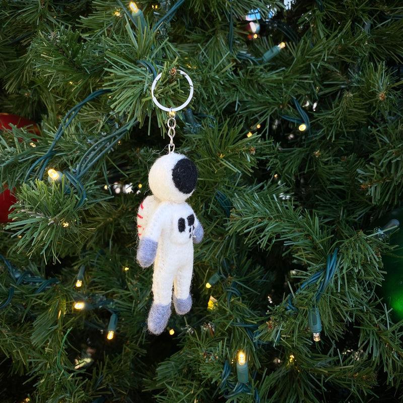 A playful wool felt astronaut keychain hangs from a branch of a Christmas tree adorned with tiny lights. The handmade felt keychain features a white spacesuit, a black visor, and gray accents on its hands and feet, adding a whimsical touch to the festive holiday decor. It is attached to a metal keychain.