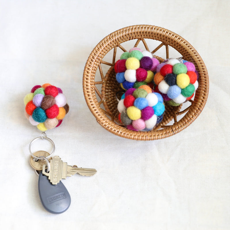 Colorful wool felt Ball Pompom keychains are displayed, with three nestled inside a small, round, light brown woven basket and one attached to keys. Each spherical felt keychain is crafted from small, vibrant wool felt balls, featuring a metal keychain with clasp. The keys include standard metal and a gray electronic key.