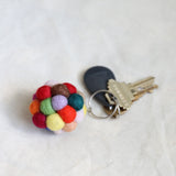 A colorful wool felt ball pompom keychain rests next to a set of keys on a textured, off-white fabric backdrop. This handmade keychain features multiple soft, fuzzy felted spheres in vibrant red, green, yellow, orange, pink, and brown, attached to a metal key ring. The handcrafted accessory evokes a playful, charming aesthetic.
