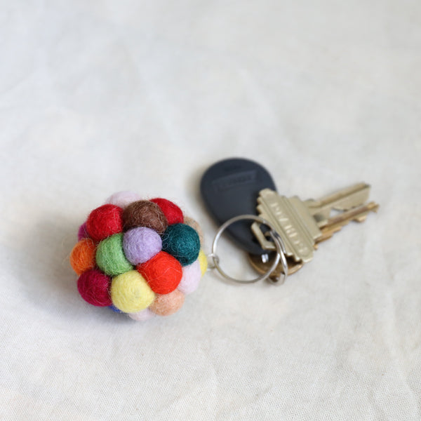 A colorful wool felt ball pompom keychain rests next to a set of keys on a textured, off-white fabric backdrop. This handmade keychain features multiple soft, fuzzy felted spheres in vibrant red, green, yellow, orange, pink, and brown, attached to a metal key ring. The handcrafted accessory evokes a playful, charming aesthetic.