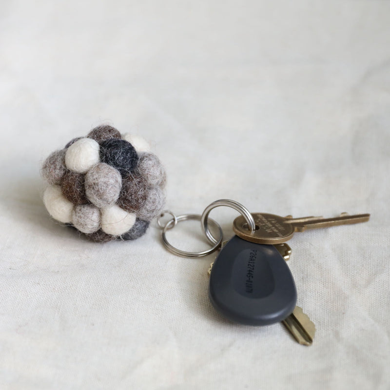 A handmade ball pompom felt keychain is displayed on a softly textured white surface, alongside a modern car key and a traditional brass key. This unique wool felt keychain features an array of small, tactile felt balls in neutral shades of white, grays, and browns. Its handcrafted appearance highlights a simple, functional aesthetic.