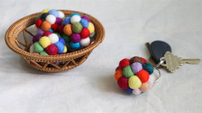 Multiple handmade Ball Pompom felt keychains are displayed in a woven basket, with one resting on a white textured surface next to a set of keys. These spherical keychains are adorned with small, colorful 100% wool felt balls in vibrant white, blue, red, and yellow, creating a tactile and cheerful design.