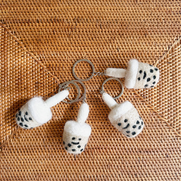 Four handmade wool felt Boba Tea keychains are displayed on a light brown woven surface, showcasing their unique charm. Each felted keychain features a rounded beige and black boba tea cup, complete with hand-stitched details, a fluffy white straw, and a silver metal key ring.