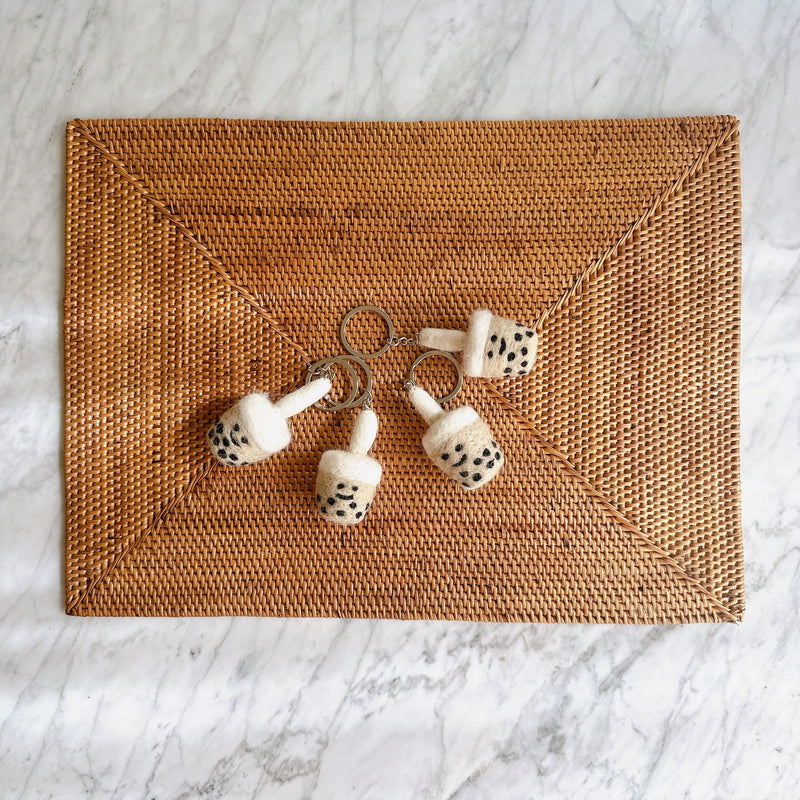 Wool felt boba tea keychains are arranged on a brown woven mat against a white marble background. Each cylindrical keychain features a white foam-like top, a tan body, and small black hand-stitched tapioca pearls with a tiny straw. A metal clasp keychain is attached to each charming felt boba tea design.