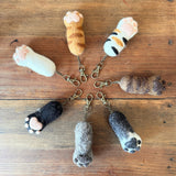 Felt cat paw keychains are arranged in a circular pattern on a wooden surface. These handmade felt keychains are 1:1 replicas of a cat's paw, showing variations in white, orange, calico, brown, black, grey, and mixed tones. Each features puffy pink toe pads and textured felt, attached to a copper keychain with clasp.