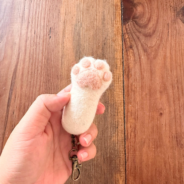 A female hand gently holds a handmade felt cat paw keychain against a rustic wooden backdrop. This unique keychain is a 1:1 replica of a cat's paw, featuring a white base with puffy, textured pink paw pads. A metal clasp is attached, emphasizing its tactile appeal and handcrafted detail.