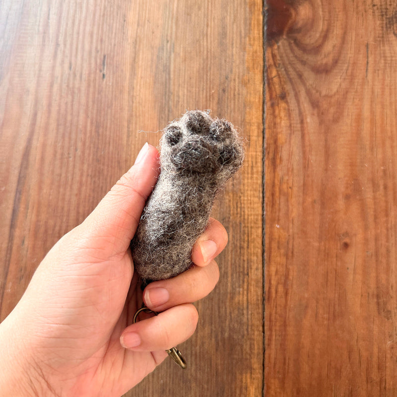 A gray wool felt cat paw keychain is gently held by a female hand against a rustic wood background. This handmade felt keychain replicates a cat's paw with detailed puffy toes. A copper keychain with a clasp dangles from the base of the paw, conveying a warm, comforting feel.