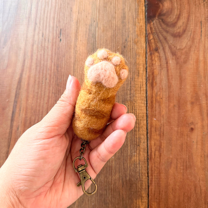 A female hand gently holds a textured orange and brown wool felt cat paw keychain against a warm wood surface. This unique wool felt keychain is shaped like a cat's paw with puffy, pink toe beans, and includes a copper keychain with a clasp. The playful design evokes a sense of comfort.