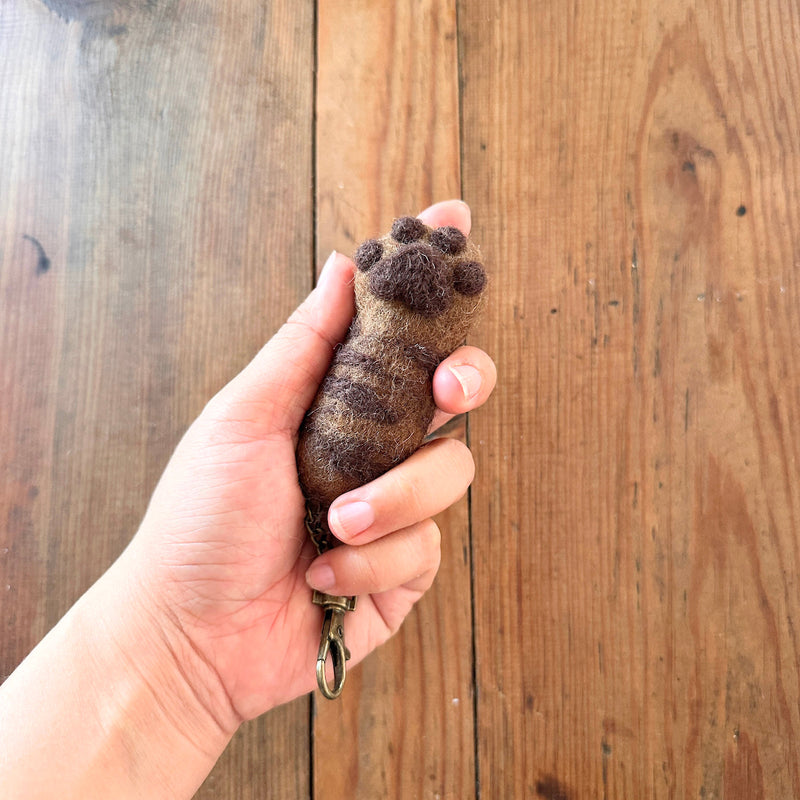 A handcrafted felt cat paw keychain is held by a female hand against a wooden background. The light brown wool felt cat paw keychain features dark brown paw pads and markings, with a textured, fur-like appearance and a copper keychain and clasp.