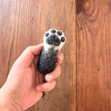 A handcrafted wool felt cat paw keychain is held by a female hand against a warm wooden background. The gray keychain features a white paw pad with small black toe beans, mimicking a real cat's paw. A copper keychain with a clasp is attached, adding to its playful and tactile appeal.