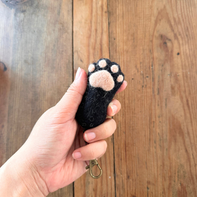 A black felt cat paw keychain is gently held by a female hand against a rustic wooden backdrop. This handmade felt keychain features a realistic cat paw design with puffy pink paw pad details. A copper keychain with a clasp is attached, completing the unique design.