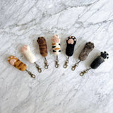 A playful row of handmade felt cat paw keychains rests on a white marble surface. These unique keychains feature various cat fur colors including white, orange, brown, black, and grey, each detailed with soft, puffy pink paw pads and attached to a copper clasp.
