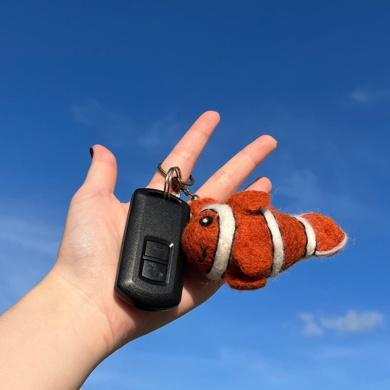 A handmade clownfish felt keychain is held by a female hand alongside a car key, set against a bright blue sky. The vibrant, high-quality plush felt clownfish features an orange body with distinctive white stripes and a small dorsal fin, attached with a metal keychain.