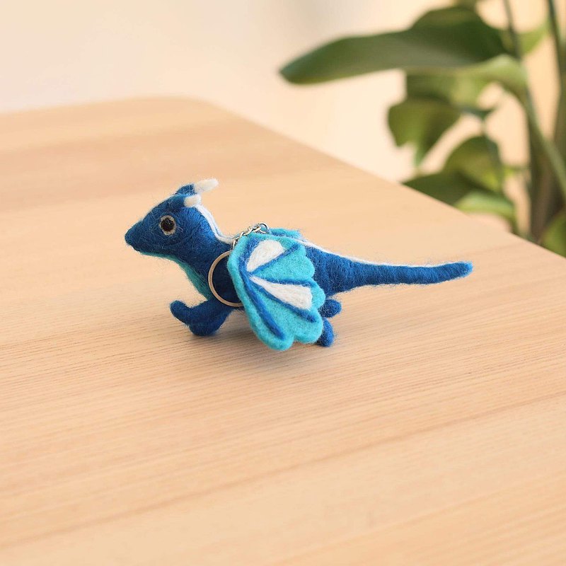 A vibrant 100% New Zealand wool felt flying dragon keychain rests on a light brown wooden surface, creating a calm, natural atmosphere. The blue felt dragon features a white horn and tail, along with two pairs of light blue and white wings. A metal clasp is attached, ready for use.