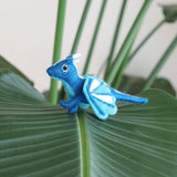 A blue wool felt flying dragon keychain rests on a vibrant green leaf against a soft white and blurred green background. This handmade felt dragon features a blue body with white horns, and wings patterned in blue and white sections. A metal keychain is attached to its back, complementing its detailed legs and long tail.