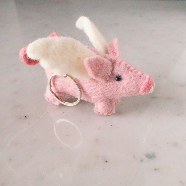 A whimsical flying pig felt keychain rests on a marbled surface in a close-up studio shot. This handmade, needle-felted wool felt keychain features a pink pig with a rounded snout, perky ears, and fluffy white wings. Attached to a metal ring, its charming design evokes a playful and lighthearted spirit.