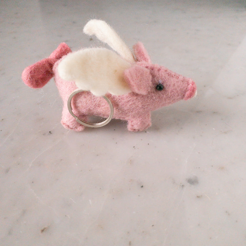A playful wool felt flying pig keychain rests on a light marble surface in a close-up studio shot. This pink handcrafted keychain features white wool felt wings, pink ears, a round snout, and a curly tail, creating an endearing and whimsical appearance. A metal keychain is attached to its body.