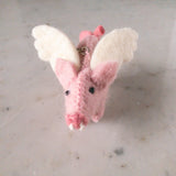 A handcrafted pink wool felt flying pig keychain is presented in a close-up shot, resting on a mottled grey and white surface. This whimsical felt keychain features white felt wings, a pink bow, black eyes, and white felt tusks, with a metal ring attached.