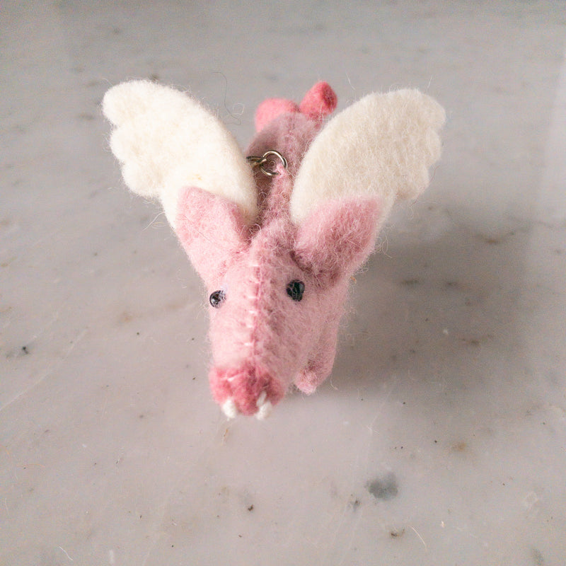 A handcrafted pink wool felt flying pig keychain is presented in a close-up shot, resting on a mottled grey and white surface. This whimsical felt keychain features white felt wings, a pink bow, black eyes, and white felt tusks, with a metal ring attached.