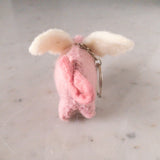 A playful wool felt flying pig keychain rests on a bright marble surface. This handcrafted pink pig features tiny wings and a soft texture. It is equipped with a metal keychain and clasp, creating a charming focal point.