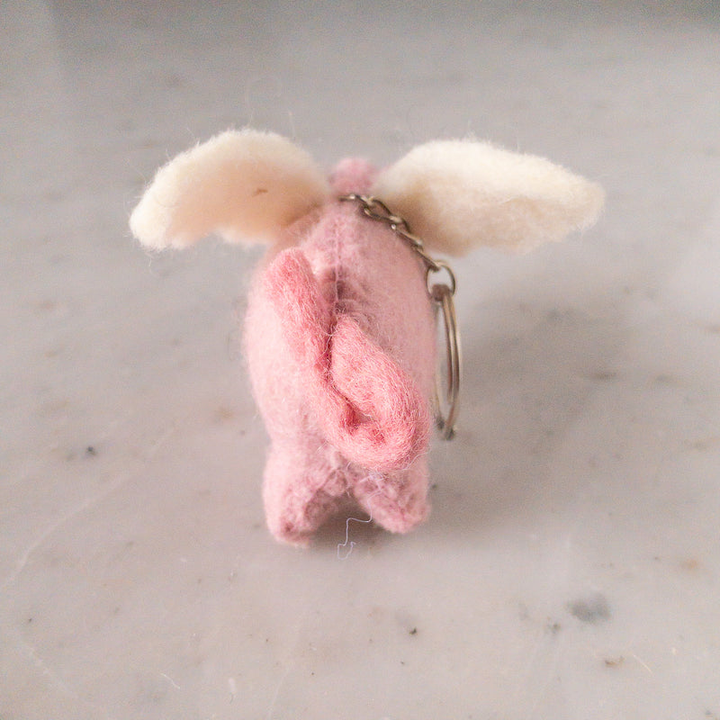 A playful wool felt flying pig keychain rests on a bright marble surface. This handcrafted pink pig features tiny wings and a soft texture. It is equipped with a metal keychain and clasp, creating a charming focal point.