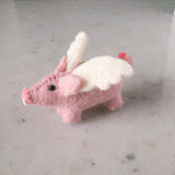 A handcrafted flying pig felt keychain is displayed against a plain background. This unique wool felt keychain is light pink, featuring white wings, a pink curly tail, and small black eyes. A copper keychain is attached near its wings. Its 100% wool material shows a clear woolen texture, highlighting its handmade charm.