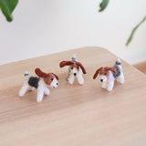 Three handmade Jack Russell wool felt keychains are playfully arranged on a light wooden surface, with a hint of greenery in the background. Each charming Jack Russell felt keychain features predominantly white wool felt with brown and black markings, floppy brown ears, a small black nose, and a short tail. All of the keychains include a metal clasp attached.
