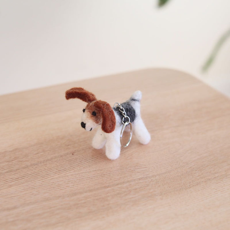A handcrafted wool felt Jack Russell keychain is displayed on a light wooden surface against a soft white backdrop. The detailed Jack Russell keychain features a white body with brown ears and head, and a black and grey back. A metal keychain with a clasp is attached to its back, adding to its functional charm.