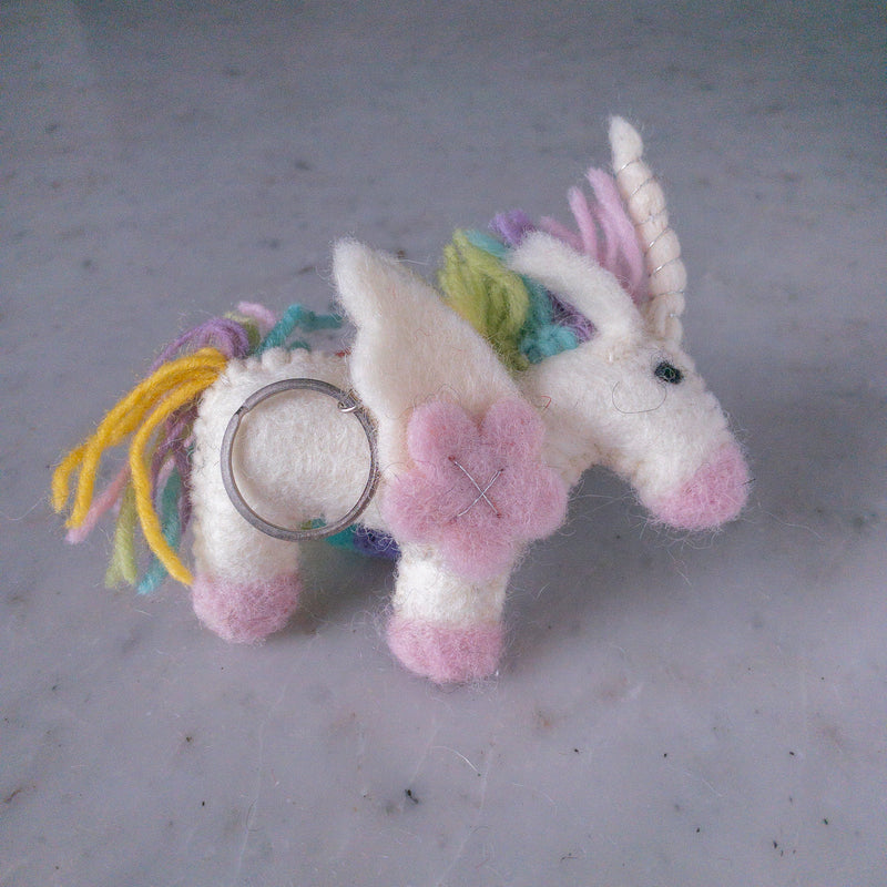 A charming wool felt pastel unicorn keychain rests on a mottled gray surface, highlighting its whimsical design. This handmade keychain features a white body with pink hooves and snout, and a playful mane with pastel yellow, green, purple, and pink strands. A silver-wrapped horn and a pink flower accent its side, completed with a metal key ring for attachment.