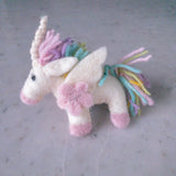 A handmade pastel unicorn felt keychain rests on a smooth, flat surface against a neutral background. This wool felt unicorn features a white body with pink hooves and muzzle. Its mane and tail display strands of pastel pink, yellow, blue, and purple, complemented by small white felt wings and a pink flower on its side. A white horn with silver thread and dark eyes complete its sweet, whimsical expression.