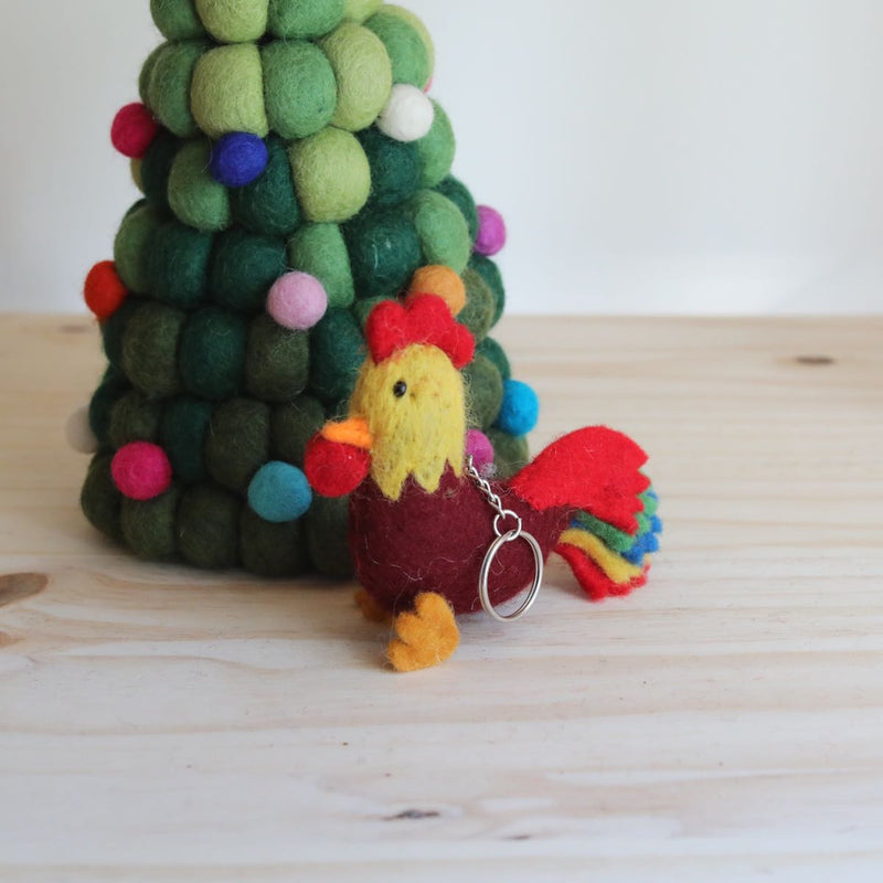 A handcrafted rooster wool felt keychain rests on a wooden surface beside a decorative felted green Christmas tree. The rooster keychain features a burgundy body, red and yellow accents, an orange beak, and vibrant red, blue, green, and yellow tail feathers, complete with a metal clasp. The festive setting includes a green felt Christmas tree adorned with small colorful felt sphere ornaments.