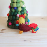 A handcrafted wool felt rooster keychain rests on a light wooden surface, positioned next to a decorative felt Christmas tree. The rooster features a burgundy body, sunny yellow neck and head, bright red crest, and a vibrant multicolored tail. This charming felted keychain stands on orange feet with an orange beak, creating a warm, festive scene.