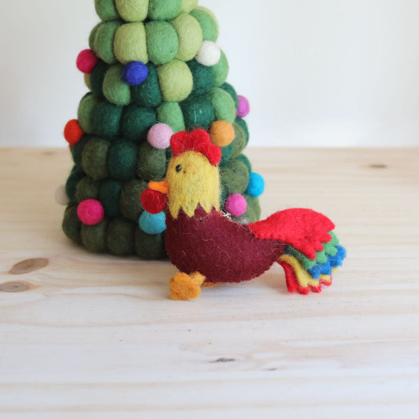 A handcrafted wool felt rooster keychain rests on a light wooden surface, positioned next to a decorative felt Christmas tree. The rooster features a burgundy body, sunny yellow neck and head, bright red crest, and a vibrant multicolored tail. This charming felted keychain stands on orange feet with an orange beak, creating a warm, festive scene.