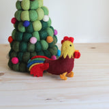 A needle felted wool rooster keychain sits on a light wooden surface, beside a felted green and colorful sphere Christmas tree, conveying a rustic, handcrafted charm. The rooster features a yellow head with a red comb and wattle, a maroon body, and a tail with red, blue, green, and yellow stripes, with yellow feet.