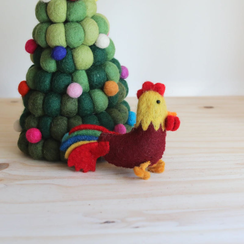A needle felted wool rooster keychain sits on a light wooden surface, beside a felted green and colorful sphere Christmas tree, conveying a rustic, handcrafted charm. The rooster features a yellow head with a red comb and wattle, a maroon body, and a tail with red, blue, green, and yellow stripes, with yellow feet.