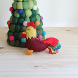 A playful wool felt rooster keychain rests on a light wooden surface. The handmade rooster features a dark red body, a yellow head with a red crest, and a vibrant tail with red, green, yellow, and blue layers. A metal clasp is attached near its neck.